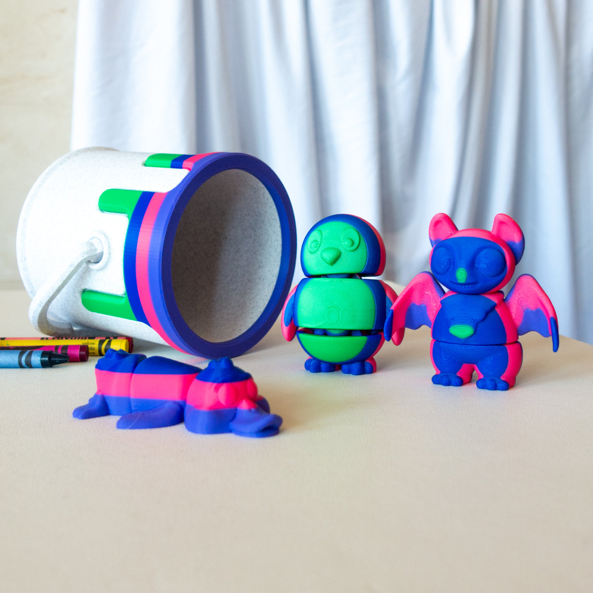 A set of colorful toy animals including a duck, a bat, and a penguin next to a paint can, displayed on a table with crayons.