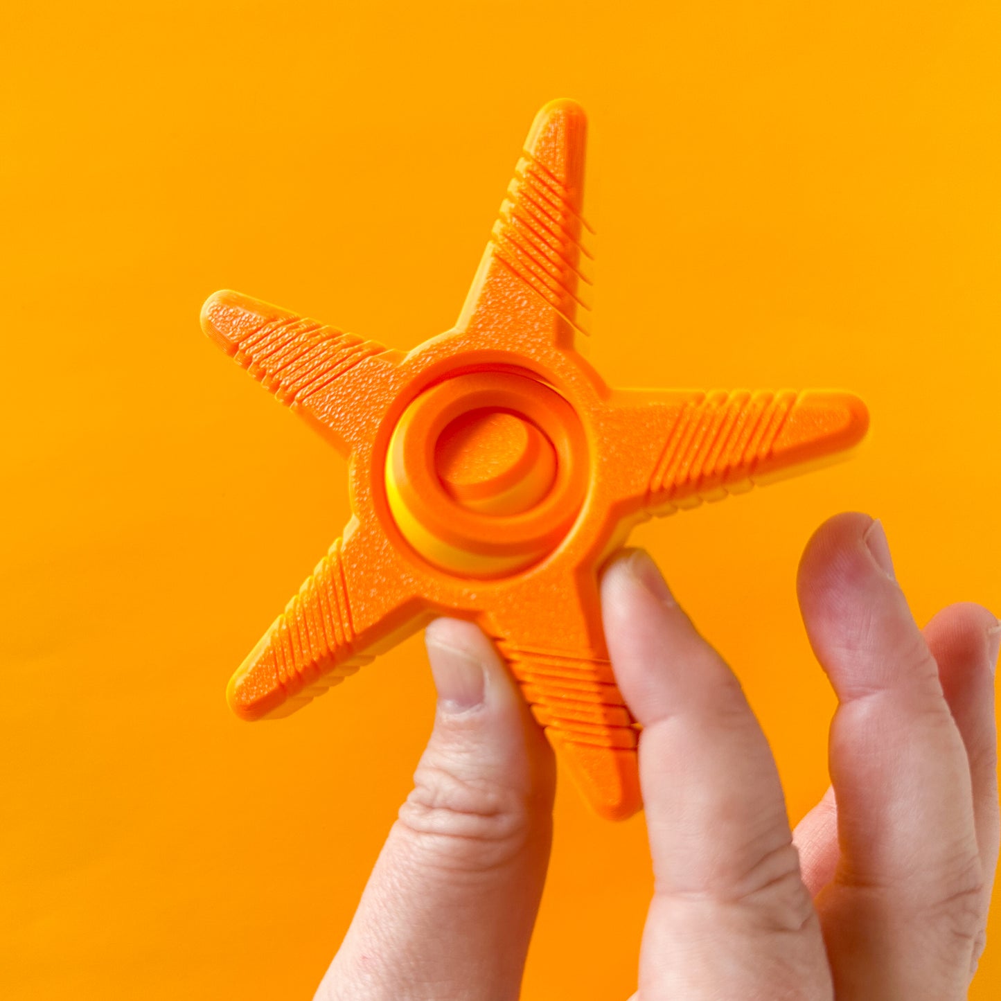 An orange and yellow five prong star spinner with a gyro center being held by a hand on a yellow backdrop